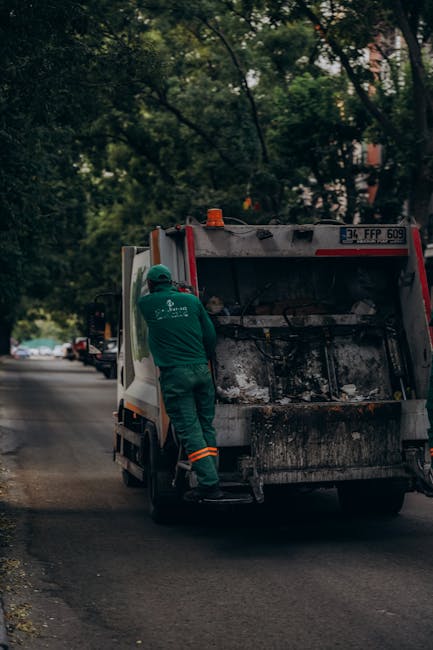 A worker wearing a green uniform and safety helmet is standing on the pavement next to an open rear loading compartment of a commercial rubbish removal truck, which is parked on a residential street l